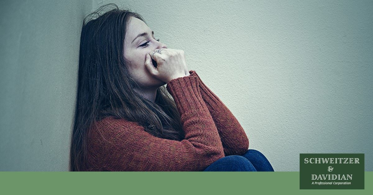 woman in red sweater sitting against wall, looking upset with hands balled up over mouth